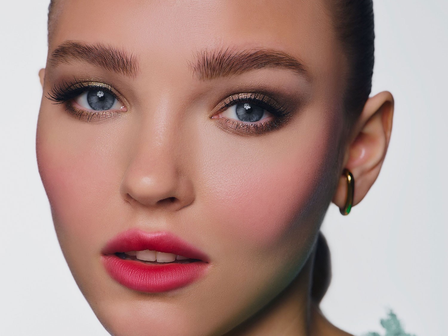 Nahaufnahme eines Frauengesichts mit strahlend blauem Blick, betont durch goldenen Lidschatten und lange Wimpern, während die Lippen in einem kräftigen Pinkton leuchten.