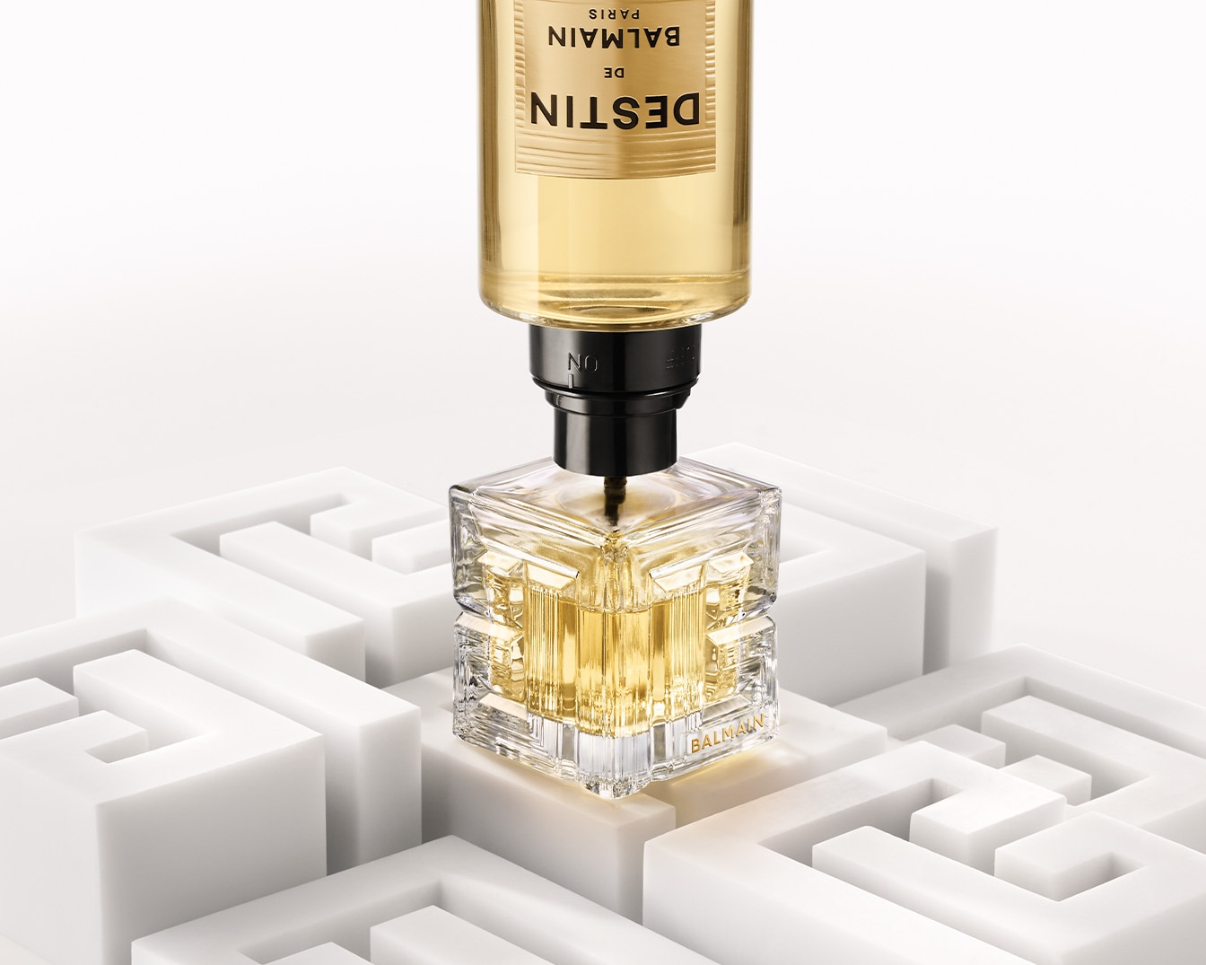 Balmain Destin perfume bottle with a clear, square design and gold liquid, set on a white maze-like structure.