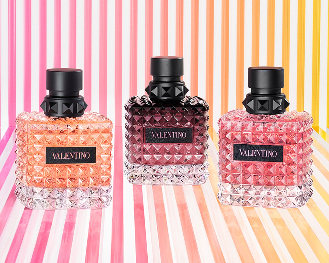 Drie Valentino geuren in levendige kleuren: oranje, bordeaux en roze, elk in een flacon met noppen en een zwarte dop, tegen een gestreepte achtergrond.