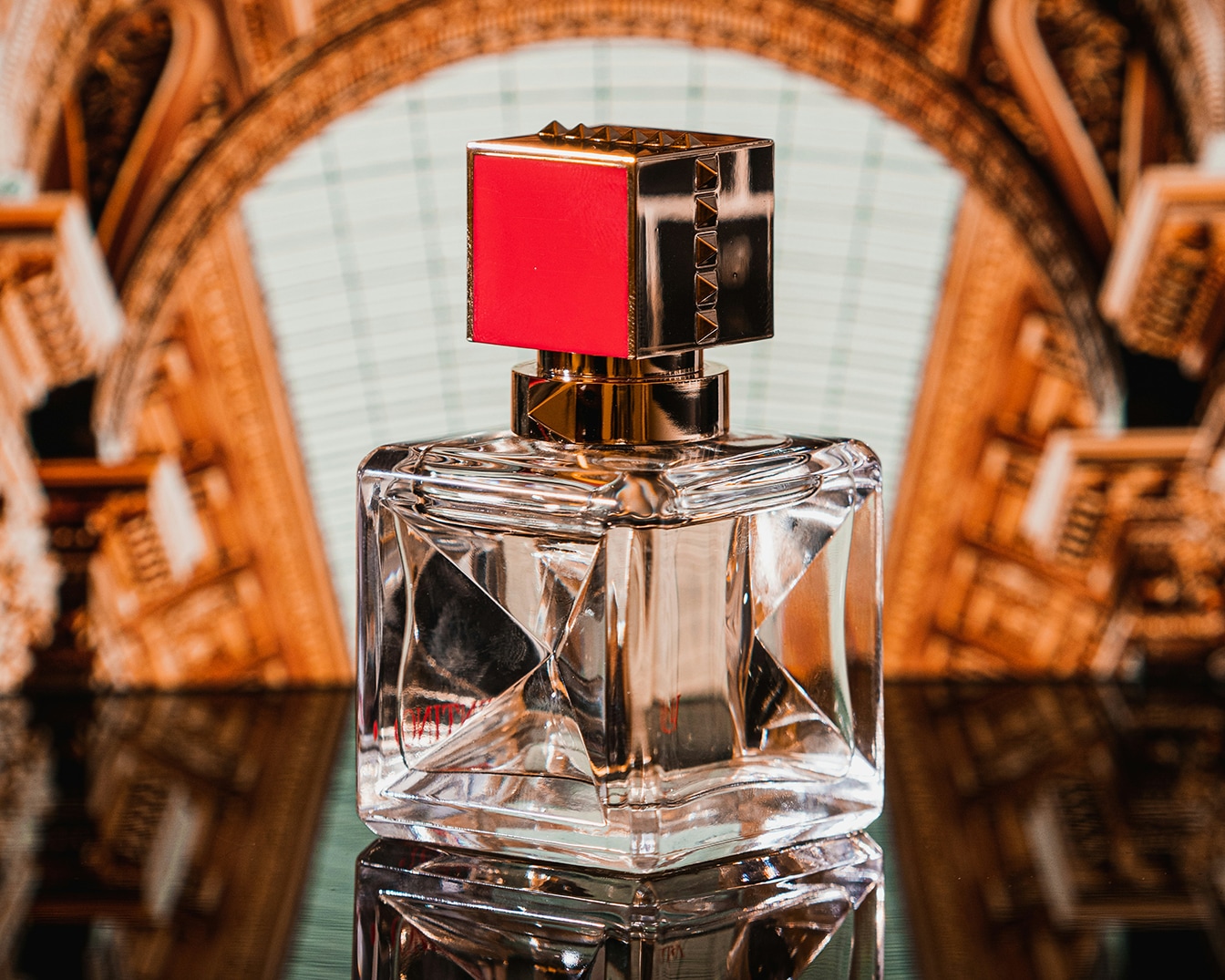 Een heldere, vierkante parfumfles met een rode en gouden dop staat op een glanzend oppervlak, klaar om je zintuigen te prikkelen.