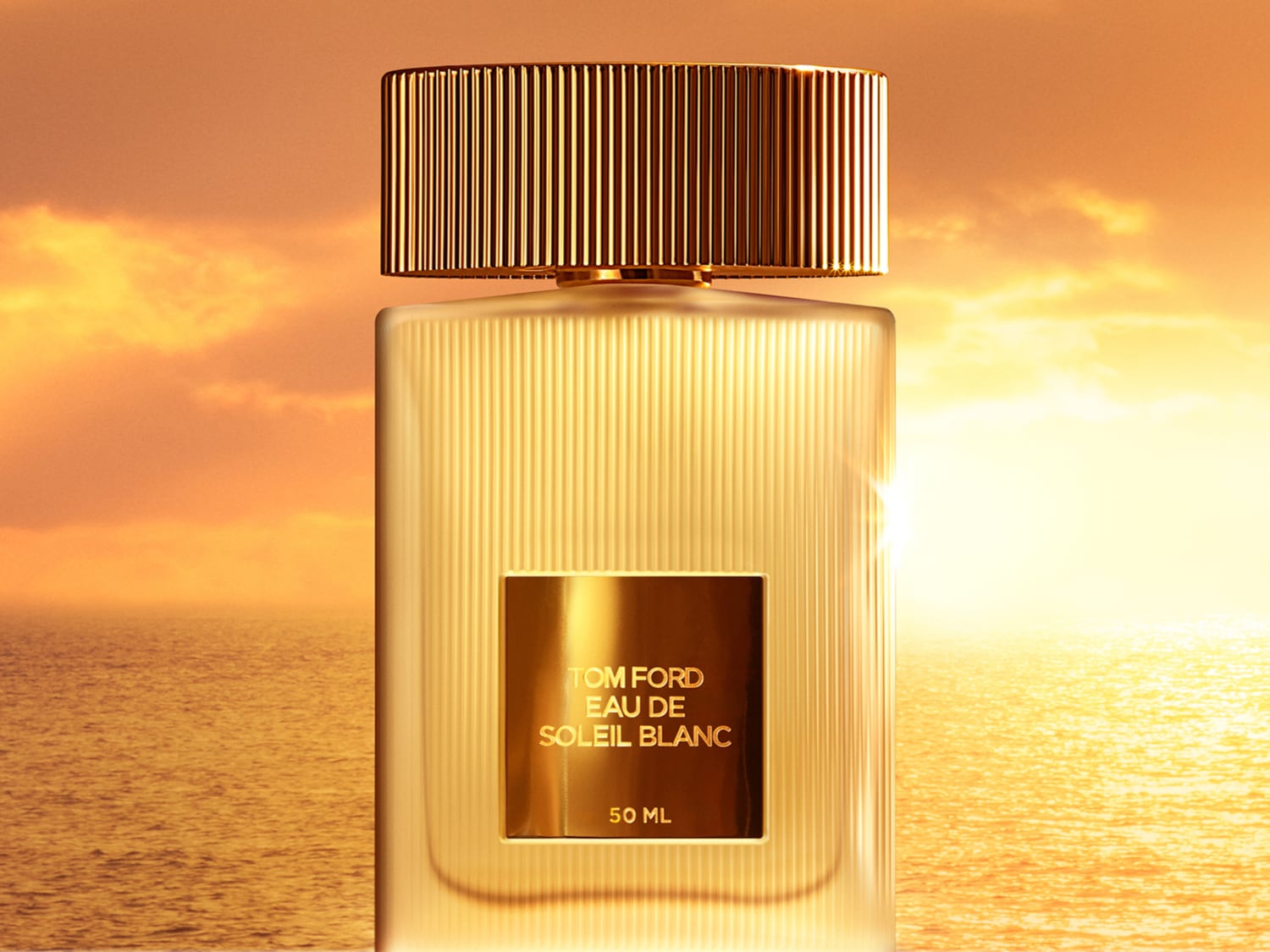 Auksinis „Tom Ford Eau de Soleil Blanc“ buteliukas priešais saulėlydžio foną virš jūros su auksiniu dangteliu ir etiketė su užrašu.