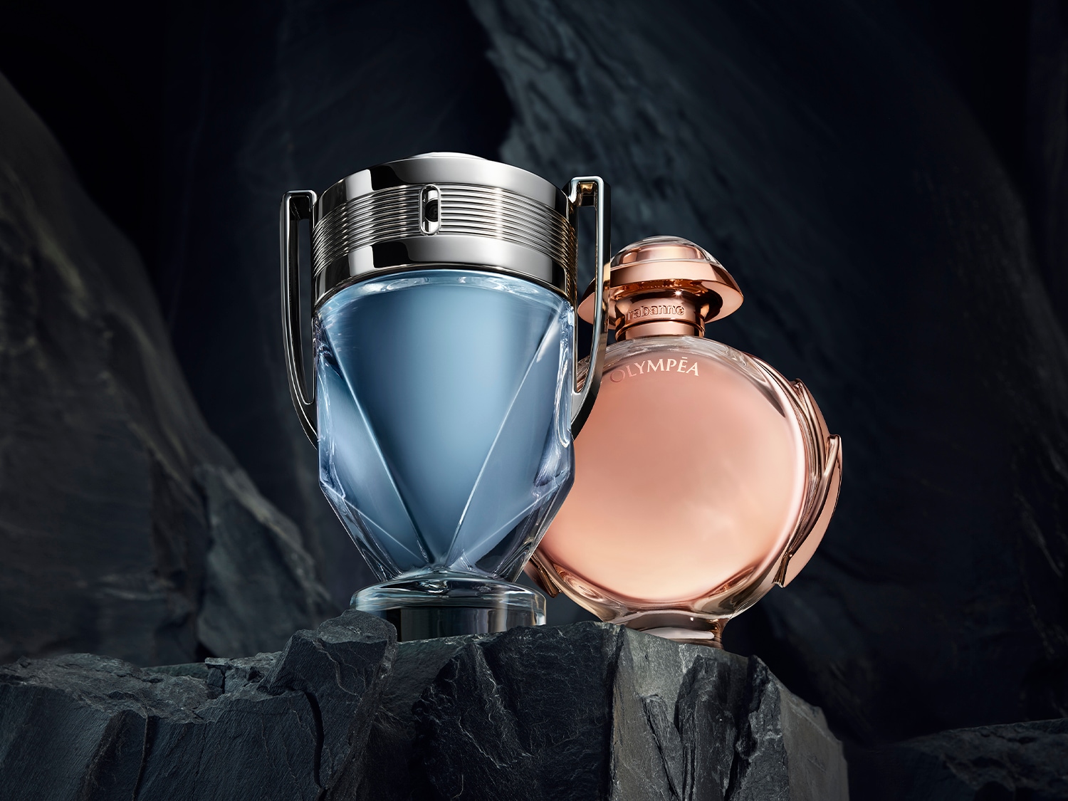Zwei Parfümflaschen, eine in Form einer silbernen Trophäe mit blauem Inhalt und die andere in einer runden, roségoldenen Flasche, stehen auf einem dunklen Felsen.