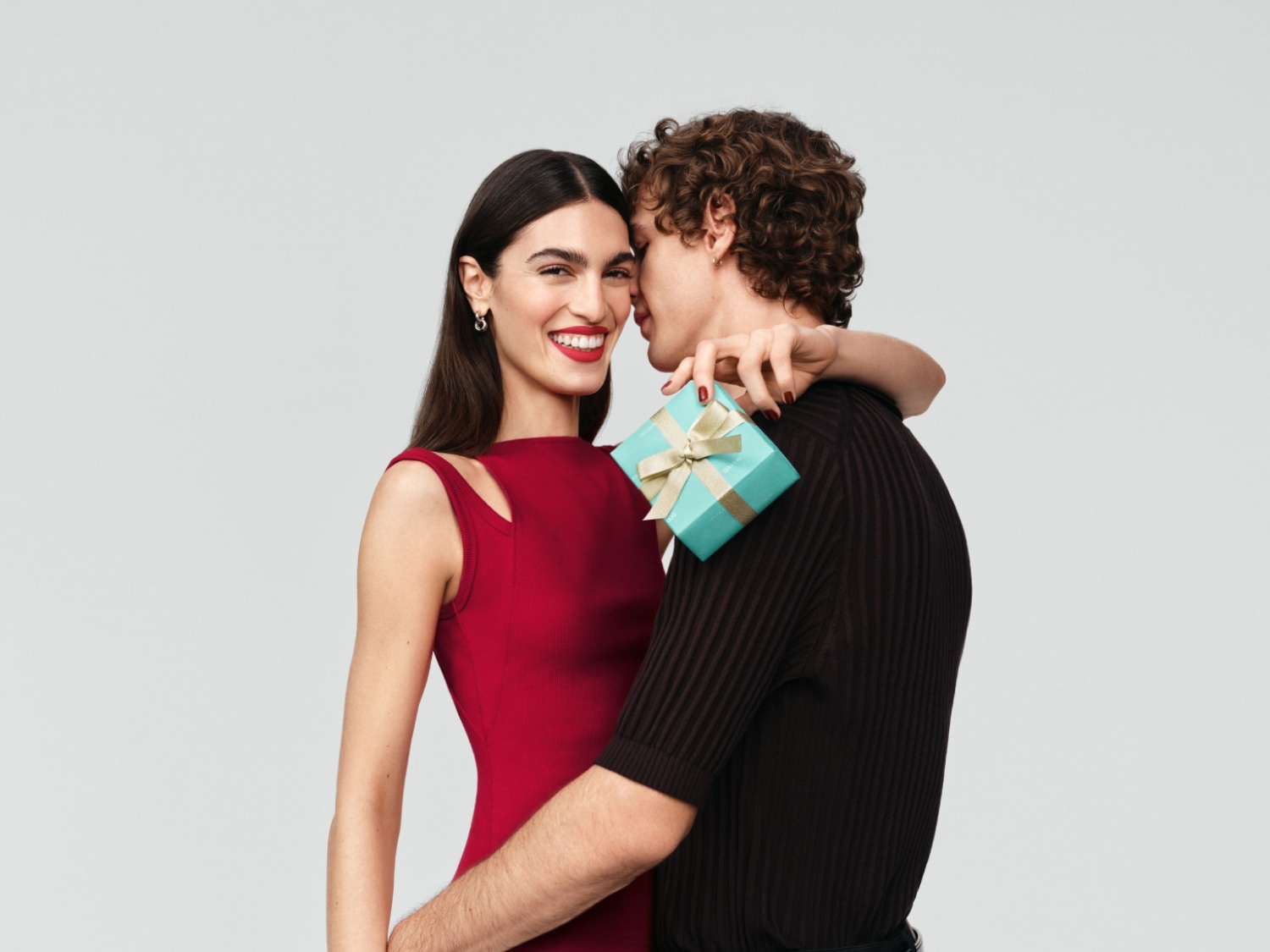 Uśmiechnięta kobieta w czerwonej sukience trzyma turkusowe pudełko z kokardą, wtulona w mężczyznę.
