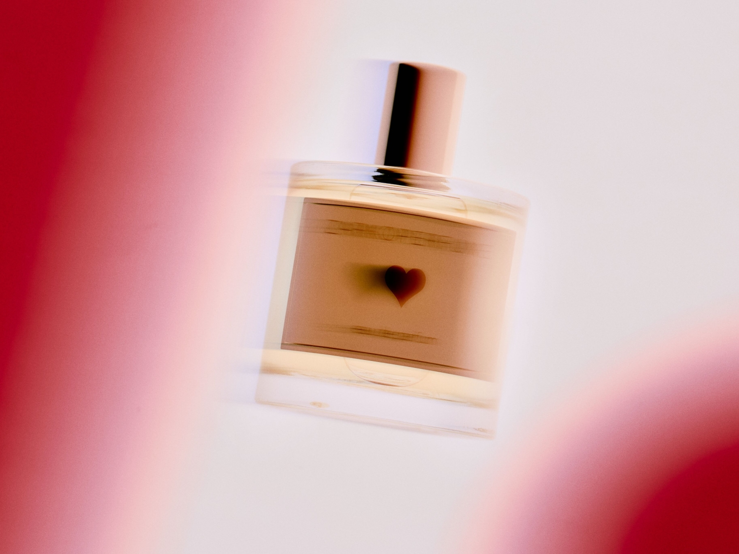 Przezroczysty flakon perfum z beżową etykietą ozdobioną małym czerwonym sercem, zamknięty złotym korkiem, na rozmytym tle w odcieniach czerwieni i bieli.