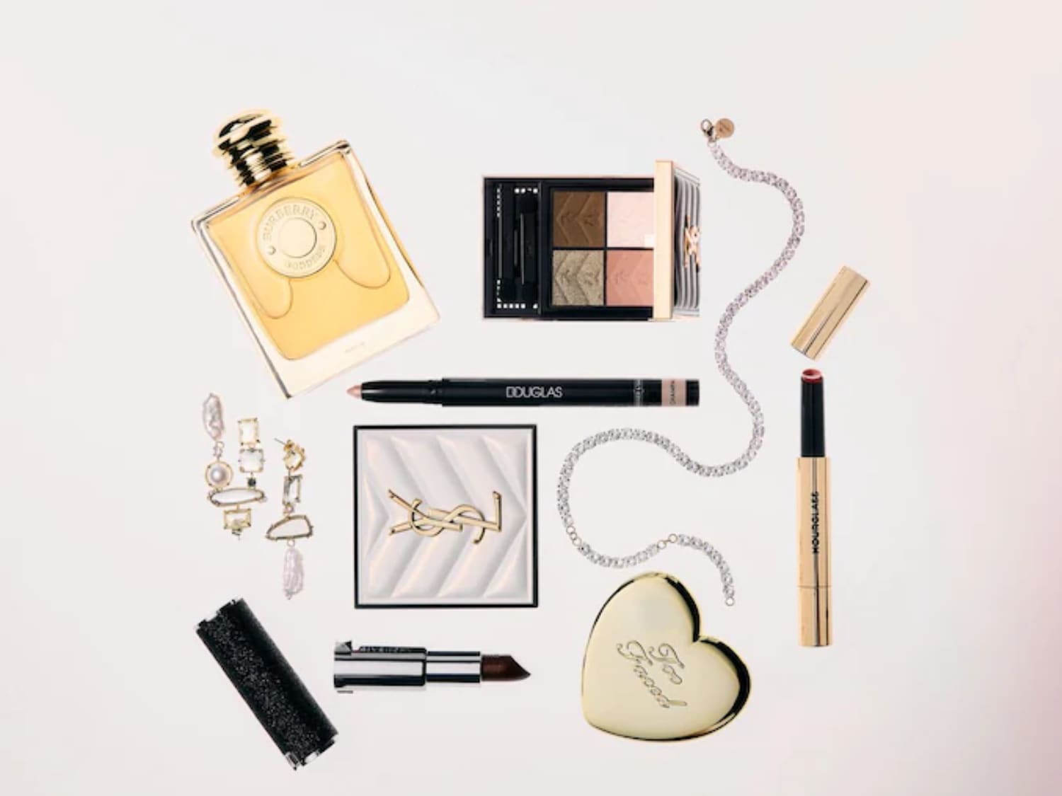 Na białym tle leżą perfumy Burberry w kwadratowej butelce z żółtym płynem, paleta cieni do powiek, kredka do oczu Douglas, rozświetlacz YSL, błyszczący czarny pilnik do paznokci, szminka Givenchy, kolczyki i łańcuszki.