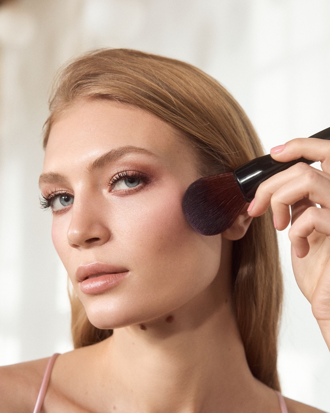 Eine blonde Frau mit hellblauen Augen trägt mit einem großen Pinsel Rouge auf ihre Wange auf, um einen frischen und strahlenden Teint zu erzielen.