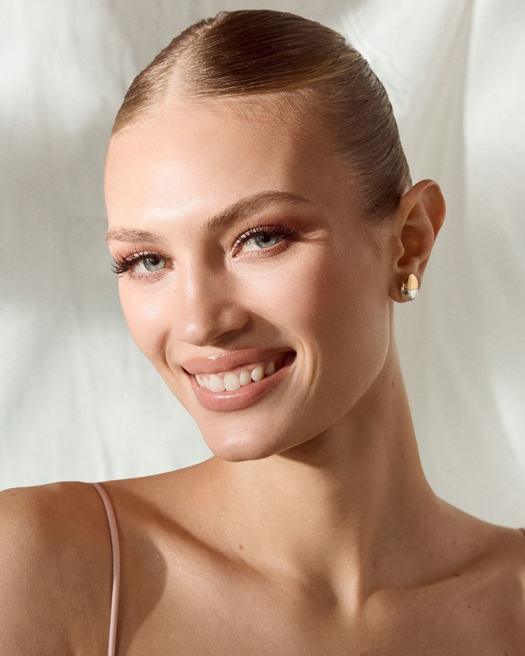 Nahaufnahme einer strahlenden Frau mit glatt zurückgekämmtem Haar, zarten rosa Lidschatten und glänzenden Lippen, die ein natürliches und frisches Aussehen verleiht.