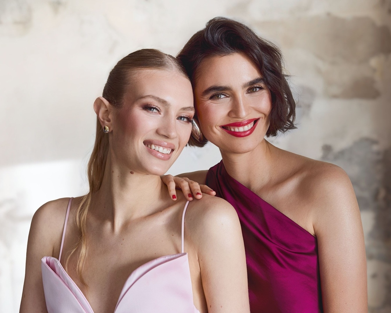 Zwei strahlende Frauen, eine in einem zarten rosa Kleid und die andere in einem eleganten, schulterfreien Kleid in dunklem Lila, lächeln selbstbewusst in die Kamera.