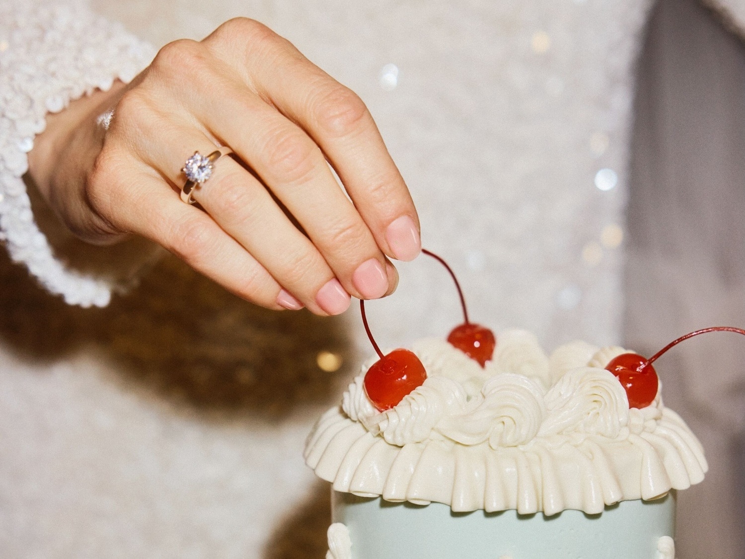 Nahaufnahme einer Hand mit einem Ring, die eine Kirsche auf einen Kuchen mit weißer Glasur und Kirschgarnierung legt.