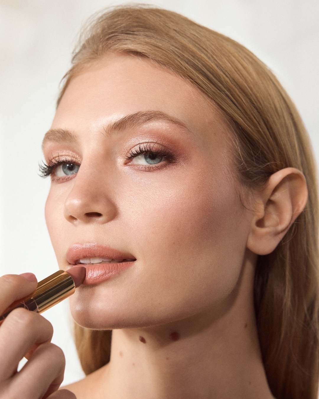 Eine blonde Frau trägt einen nudefarbenen Lippenstift auf, der in einer goldenen Hülse steckt, und ihr Make-up ist in warmen Rosatönen gehalten.