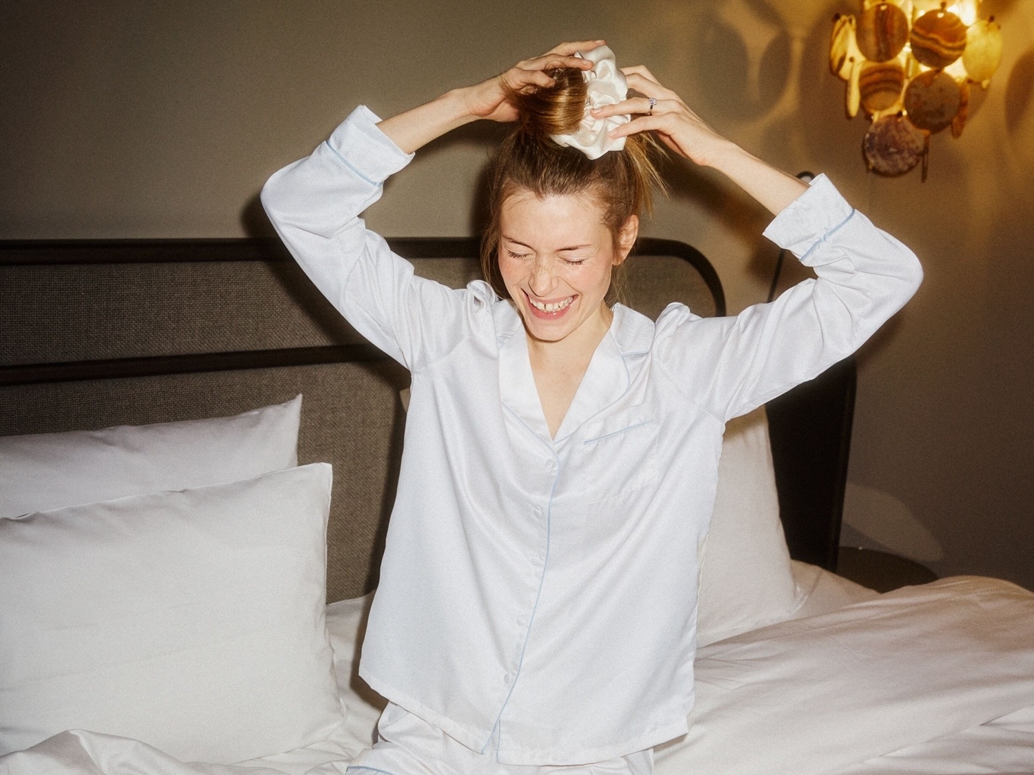 Eine lächelnde Frau im weißen Pyjama sitzt auf einem Bett und bindet ihr Haar mit einem weißen Scrunchie zusammen.