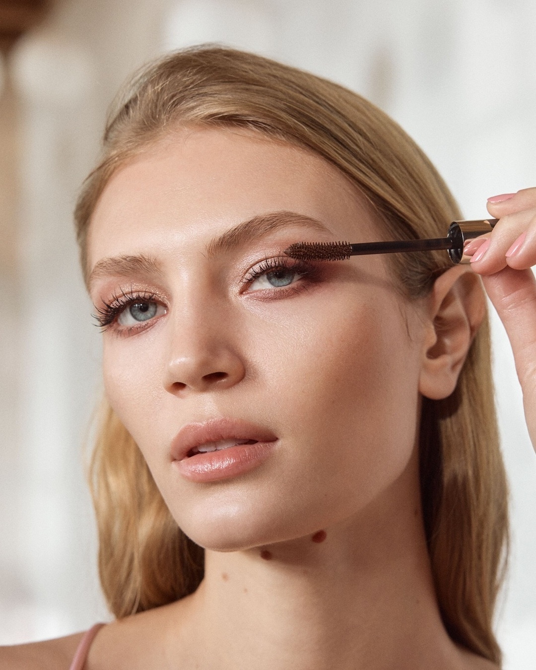 Eine blonde Frau mit blauen Augen trägt braune Mascara mit einer goldenen Bürste auf ihre oberen Wimpern auf.