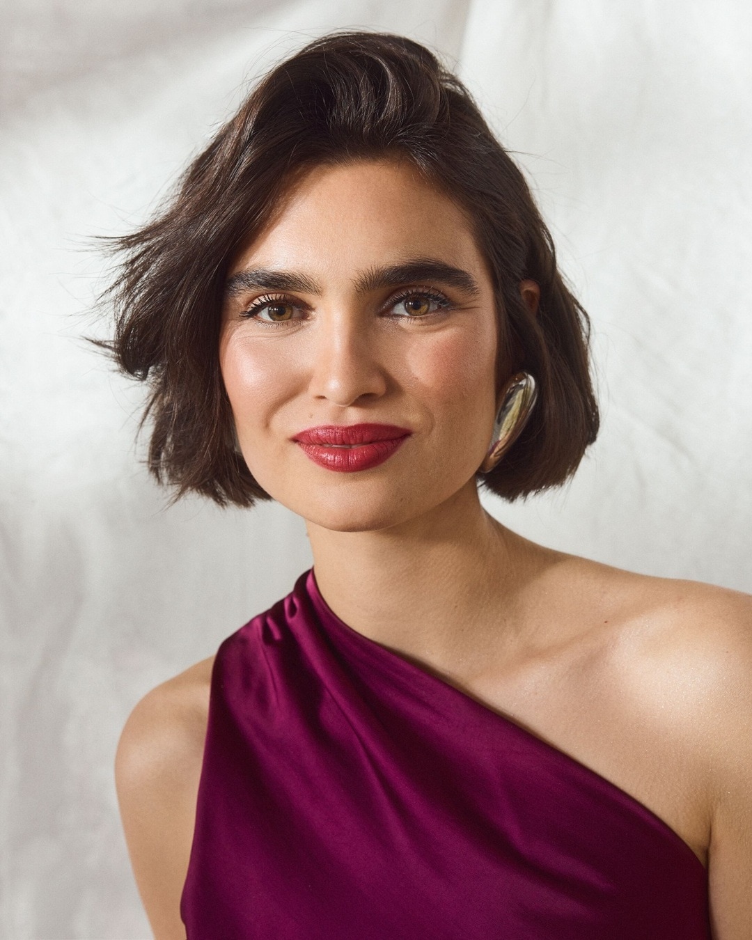 Porträt einer Frau mit braunen, gewellten Haaren, roten Lippen und einem violetten, schulterfreien Kleid, die einen silbernen Ohrring trägt und in die Kamera lächelt.