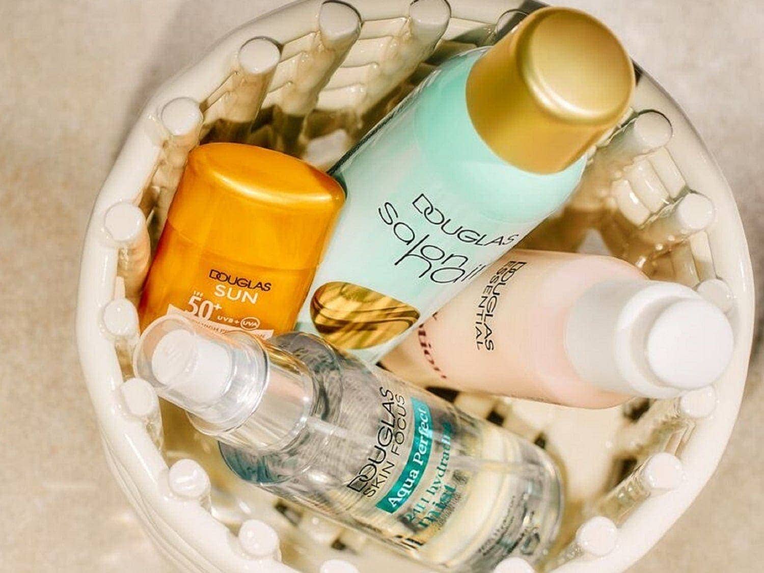 In einem weißen Korb liegen verschiedene Douglas-Produkte, darunter Sonnenschutzmittel in einer orangefarbenen Flasche, Aqua Perfect 2in1 Hydrat-Nebel und Salobil.