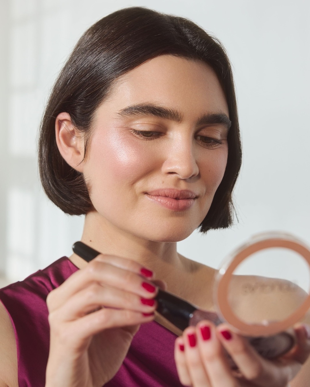 Eine Frau mit kurzen, dunklen Haaren und rotem Nagellack trägt mit einem Pinsel Bronzepuder auf ihr Gesicht auf.