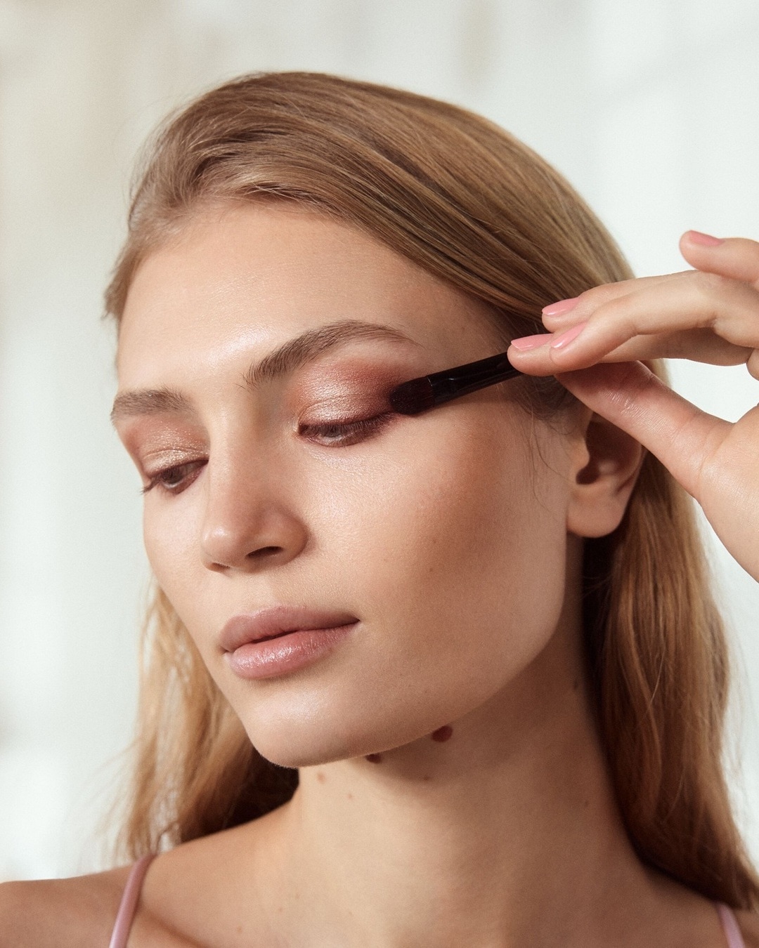 Eine Frau mit blonden Haaren trägt mit einem Pinsel schimmernden, rosafarbenen Lidschatten auf ihr Augenlid auf.