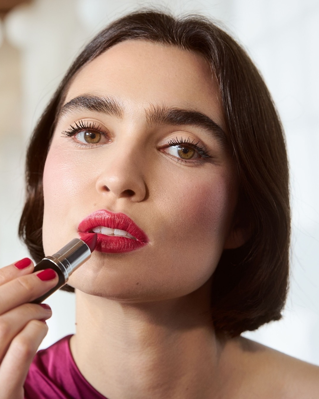 Eine Frau mit braunen Haaren und roten Nägeln trägt roten Lippenstift auf, um ihren Look zu vervollständigen.