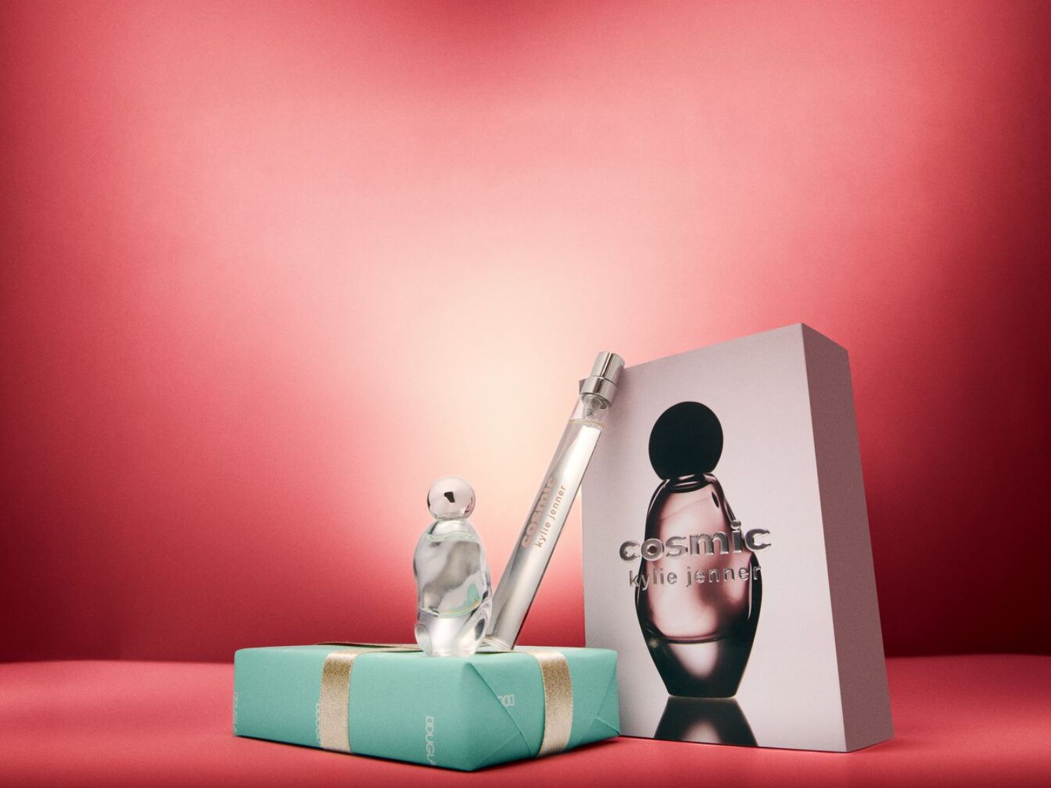 Kosmická vůně Kylie Jenner je prezentována s miniaturou, cestovním sprejem a balením na pozadí s červeným gradientem.