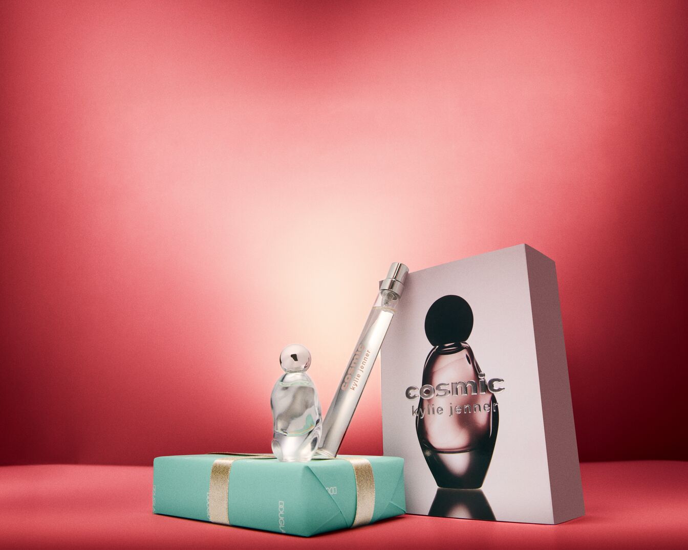A Cosmic Kylie Jenner parfümcsalád termékei láthatók: egy kis üveg parfüm egy türkizkék ajándékdobozon, egy utazó méretű parfüm és a parfüm doboza.