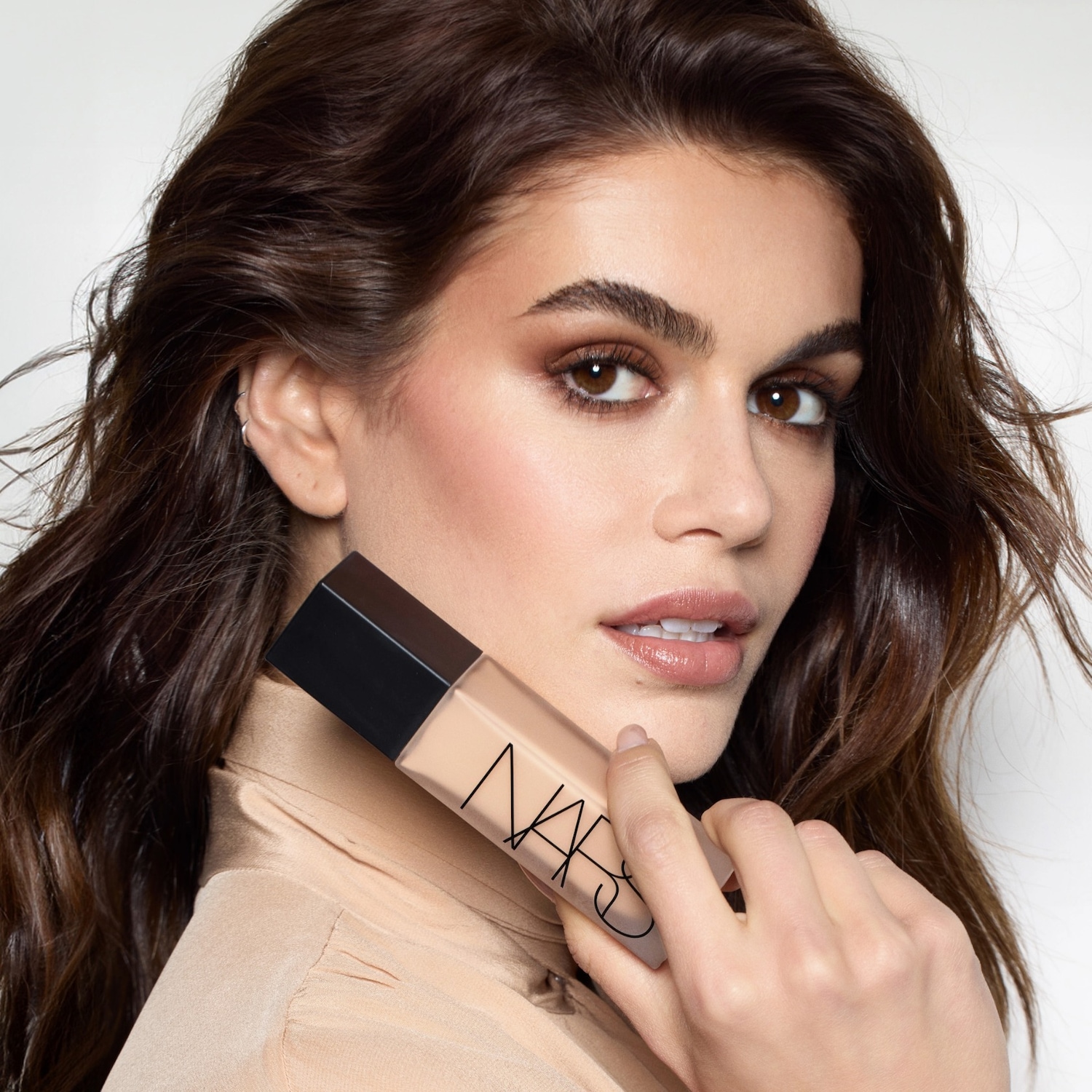 Une femme aux longs cheveux bruns tient un fond de teint NARS beige dans un flacon rectangulaire avec un bouchon noir, mettant en valeur un teint impeccable et un maquillage naturel.