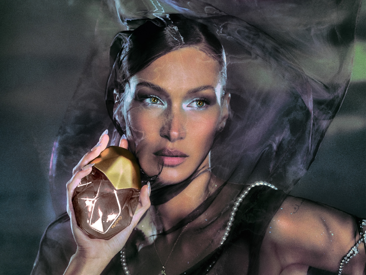 Une femme tient un flacon de parfum Orabela en forme de pierre précieuse avec un bouchon doré, enveloppée d'un voile noir transparent, créant une ambiance mystérieuse et élégante.