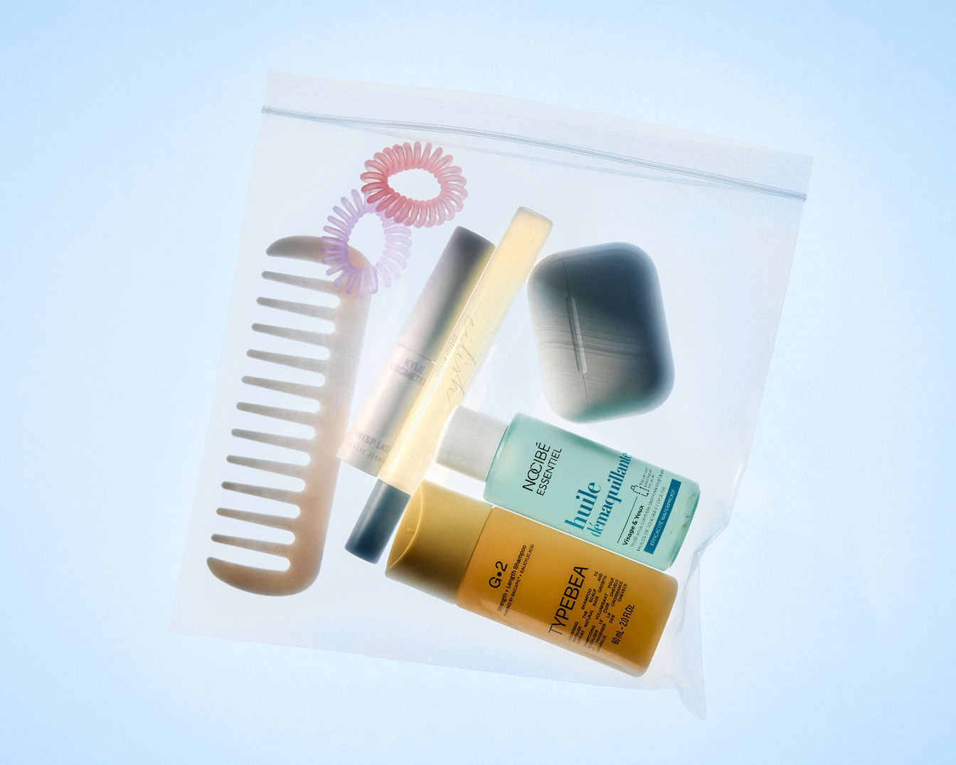 Dans une trousse transparente, on trouve un peigne beige, des élastiques à cheveux roses, un mascara Kylie Cosmetics, un stick correcteur, un beauty blender gris, une huile démaquillante Nocibé Essentiel et un shampoing Typebea.