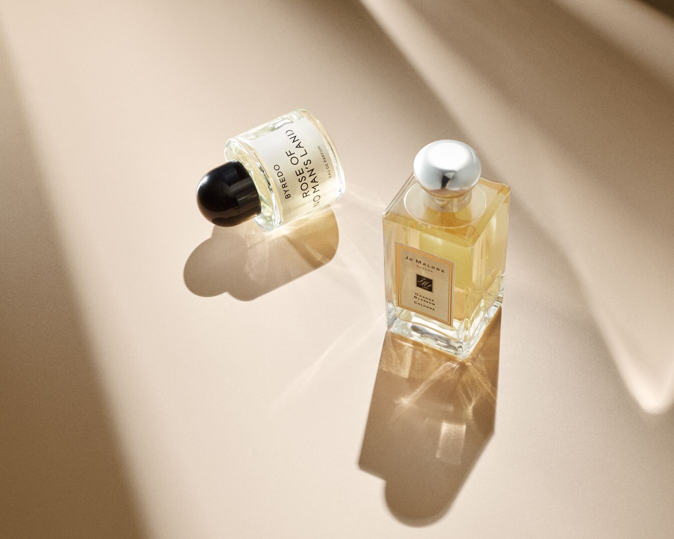Zwei Parfümflaschen, eine von Byredo Rose of No Man's Land mit schwarzem Verschluss und eine von Jo Malone Orange Blossom Cologne mit silbernem Verschluss, stehen auf einem beigen Untergrund.