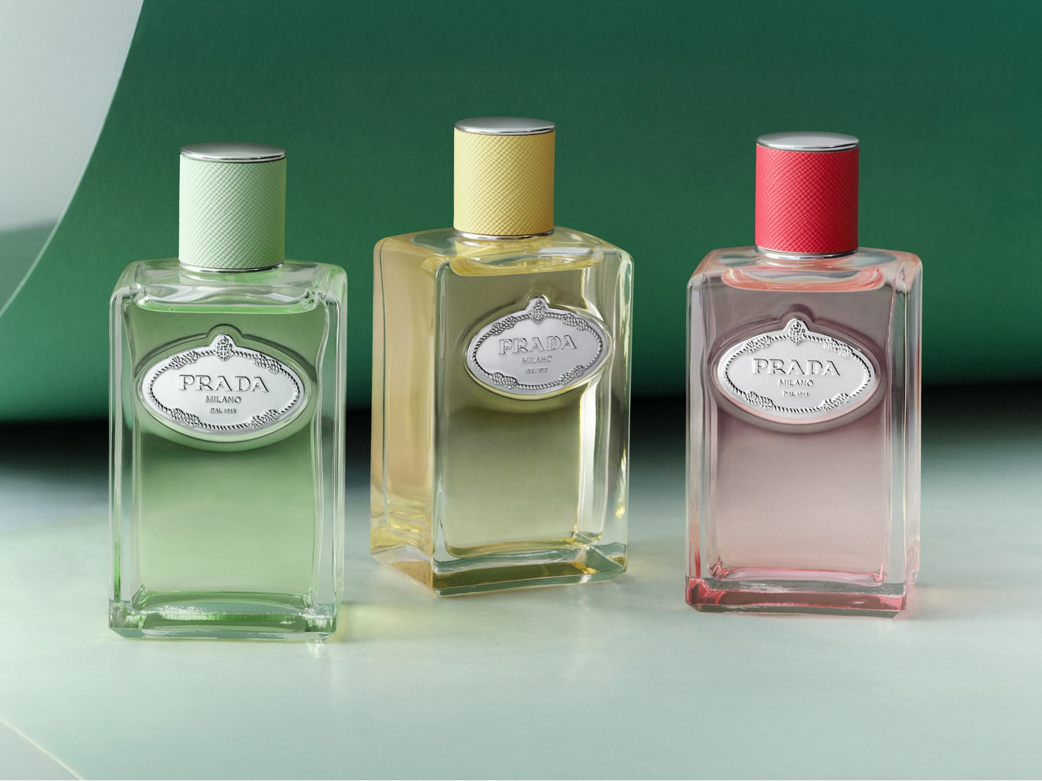 Tres frascos de perfume Prada Milano, cada uno con un líquido de color diferente: verde, amarillo y rosa, con tapas a juego y una etiqueta plateada.