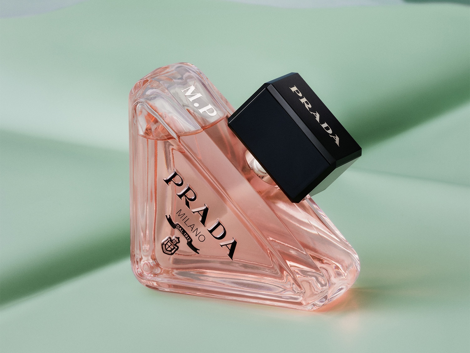 El frasco de perfume Prada, con su líquido rosado y tapón negro, irradia elegancia y sofisticación sobre un fondo verde suave.