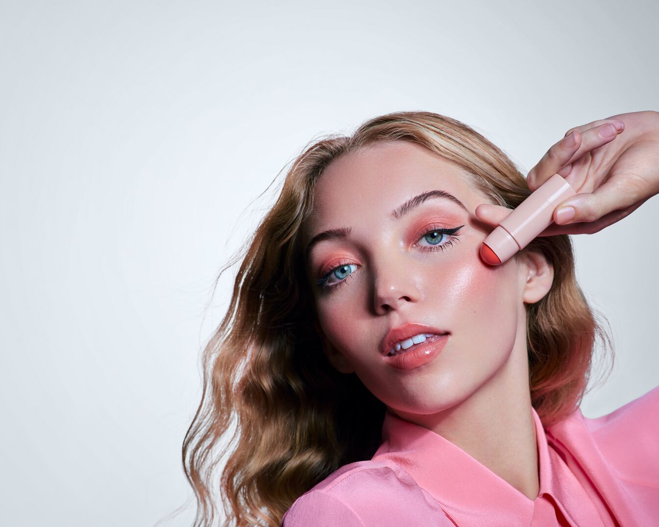 Une femme blonde aux yeux bleus applique un blush crème rose sur sa joue avec un stick à l'extrémité rouge, pour un teint frais et lumineux.