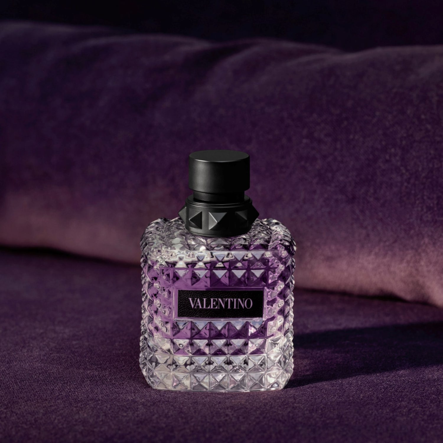 Nahaufnahme eines Valentino-Parfums in einem durchsichtigen, facettierten Flakon mit schwarzem Verschluss und Etikett, das auf einem violetten Samtstoff steht.