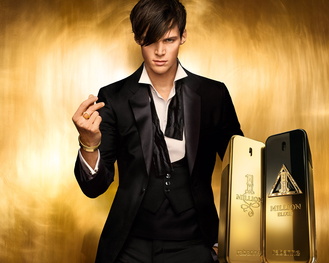 Un homme élégant en smoking noir, orné de bijoux dorés, se tient devant deux flacons de parfum "1 Million Elixir" de Rabanne, l'un doré et l'autre noir, sur un fond doré scintillant.