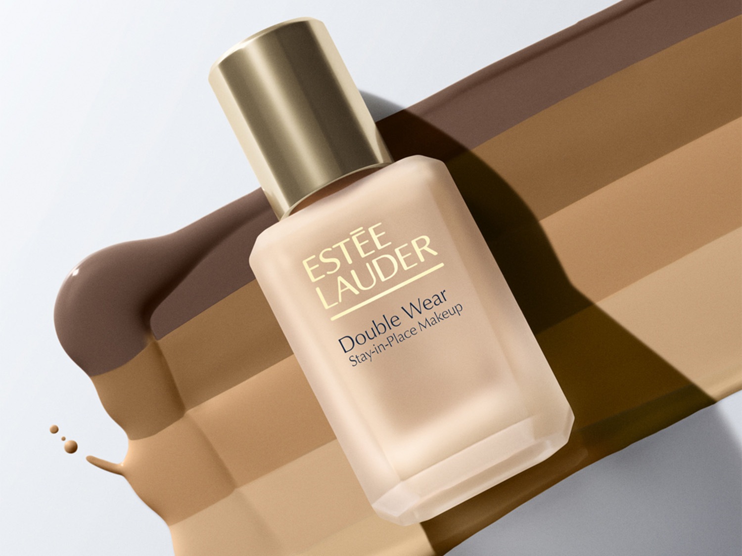 Découvrez le fond de teint Double Wear Stay-in-Place d'Estée Lauder dans un flacon en verre dépoli avec un bouchon doré, présenté sur des échantillons de différentes nuances de beige à marron.