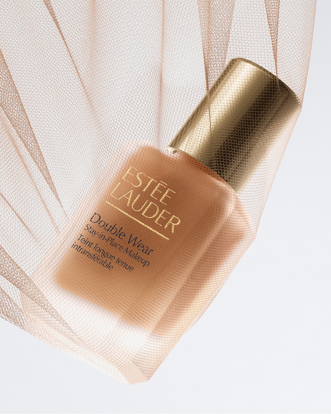 Découvrez le fond de teint Estée Lauder Double Wear Stay-in-Place Makeup, un teint longue tenue intransférable, présenté dans un flacon élégant avec un bouchon doré, le tout enveloppé dans un voile délicat.
