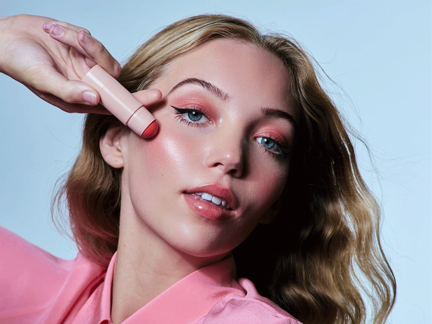 Eine Frau mit blonden, gewellten Haaren und einem rosa Hemd trägt einen Rouge-Stick auf ihre Wange auf, um einen frischen, rosigen Teint zu erzielen.