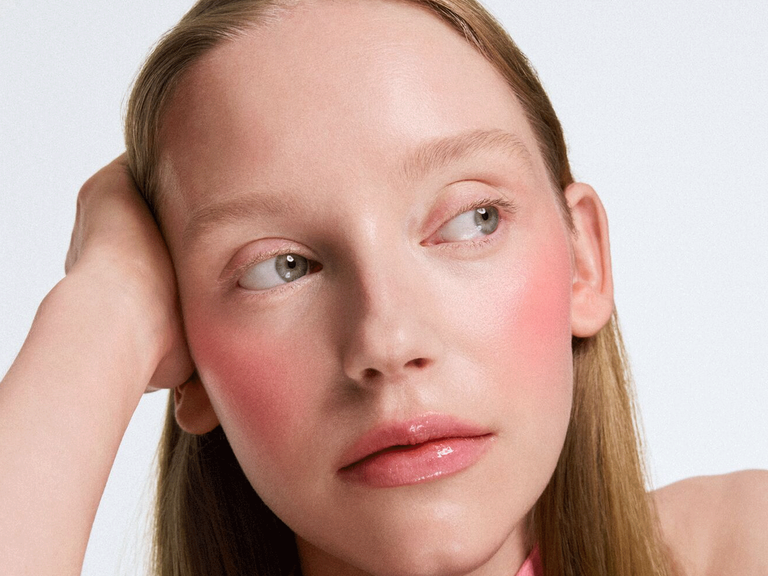 Nahaufnahme eines hellhäutigen Models mit rosa Rouge und glänzenden Lippen, das mit dem Kopf auf der Hand ruht und zur Seite blickt.