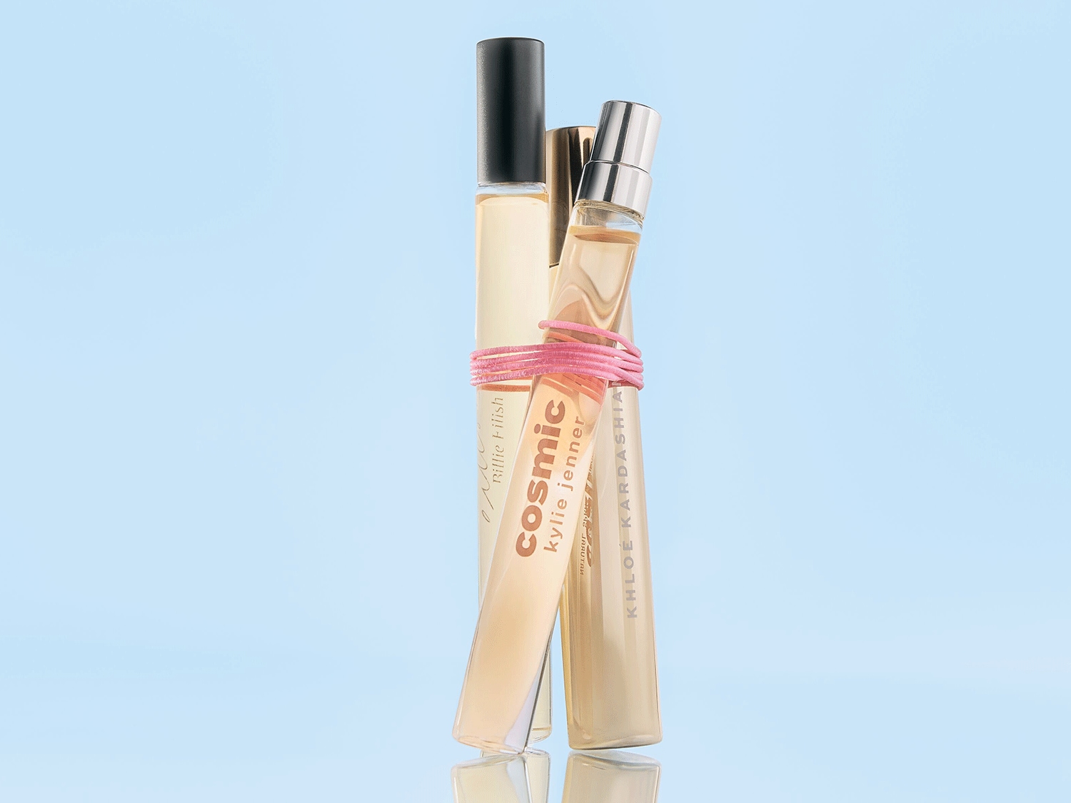 Nahaufnahme von drei Parfümflaschen, die mit einem rosa Gummiband zusammengebunden sind, darunter Billie Eilish, Cosmic Kylie Jenner und Khloe Kardashian, vor einem hellblauen Hintergrund.