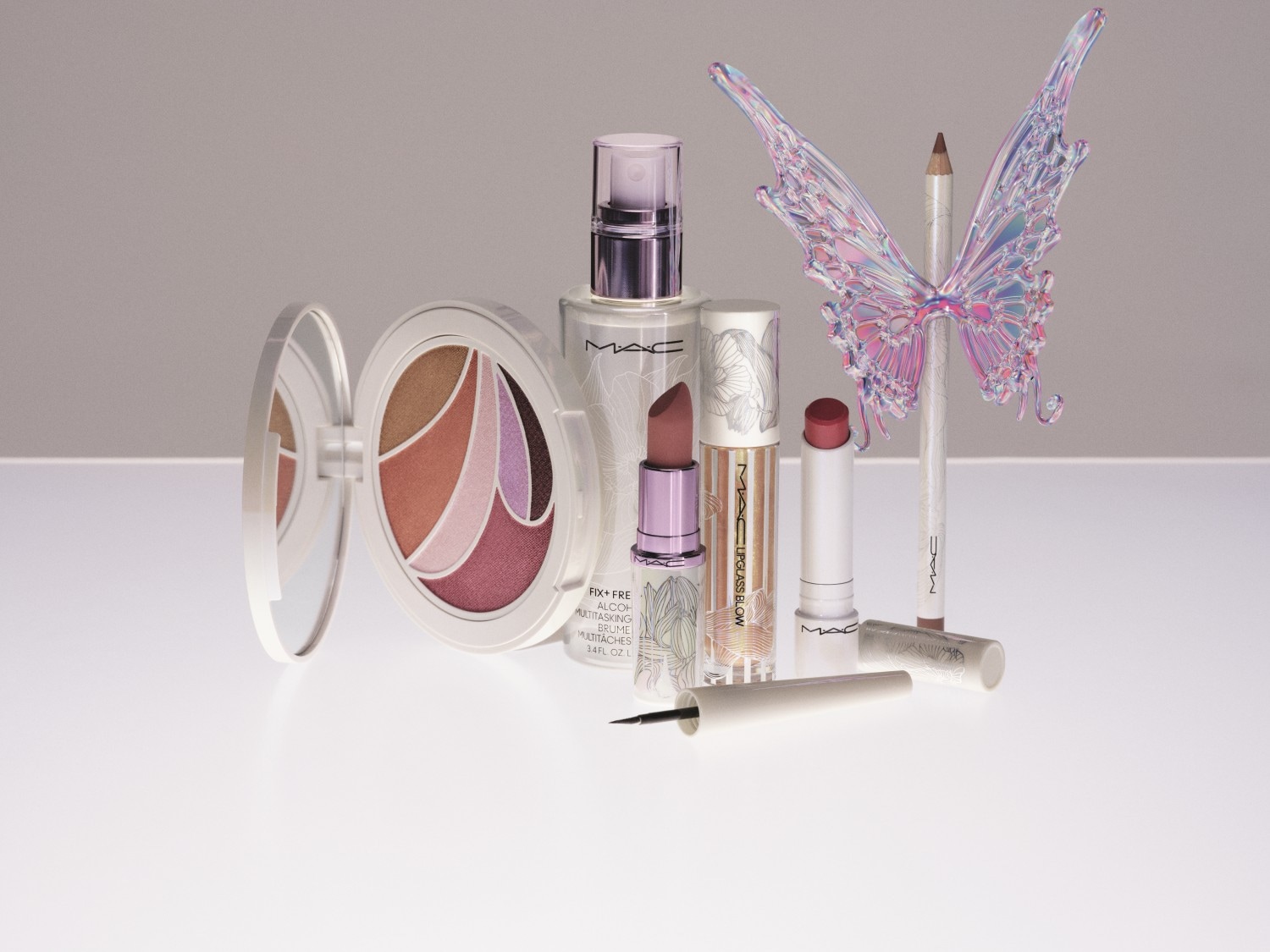 Paleta cieni do powiek MAC w okrągłym, białym opakowaniu z lusterkiem obok mgiełki utrwalającej makijaż, błyszczyka, szminki, kredki do oczu i ozdobnego motyla.