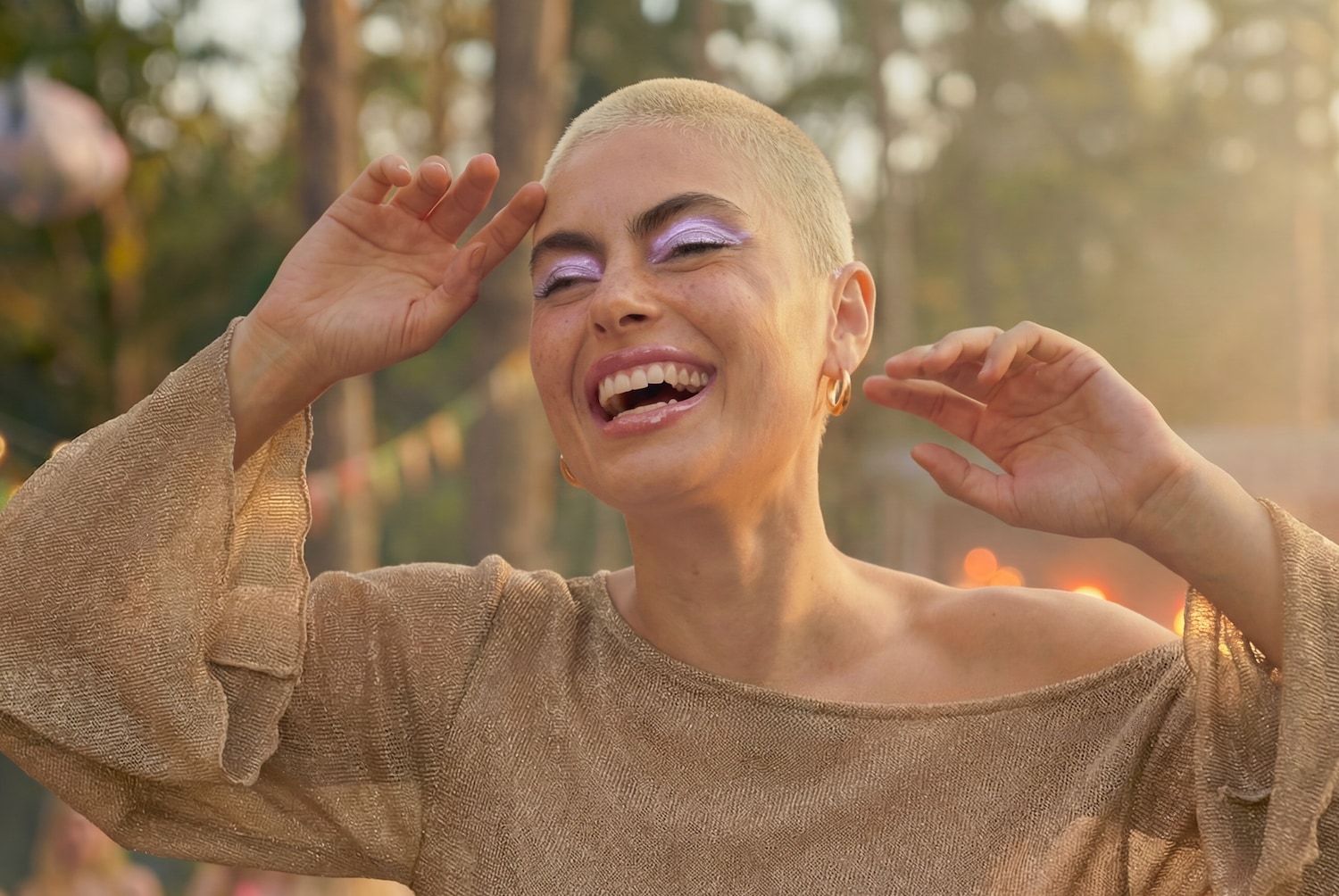 Eine strahlende Frau mit kurz geschorenem Haar und goldenen Creolen lacht, ihr lila Lidschatten funkelt im Sonnenlicht.
