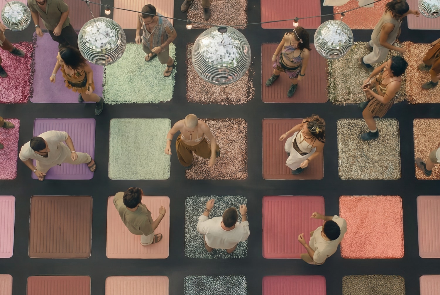 Von oben zeigt das Bild eine Gruppe von Menschen, die auf quadratischen Plattformen in verschiedenen Farben tanzen, die an eine Lidschattenpalette erinnern, während Discokugeln von der Decke hängen.