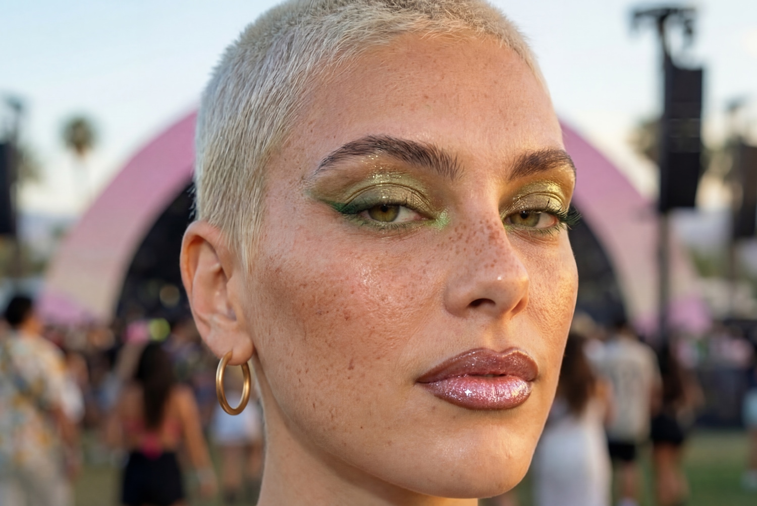 Nahaufnahme eines Gesichts mit schimmerndem grünen Lidschatten, glänzenden Lippen und goldenen Ohrringen vor einem verschwommenen Hintergrund.