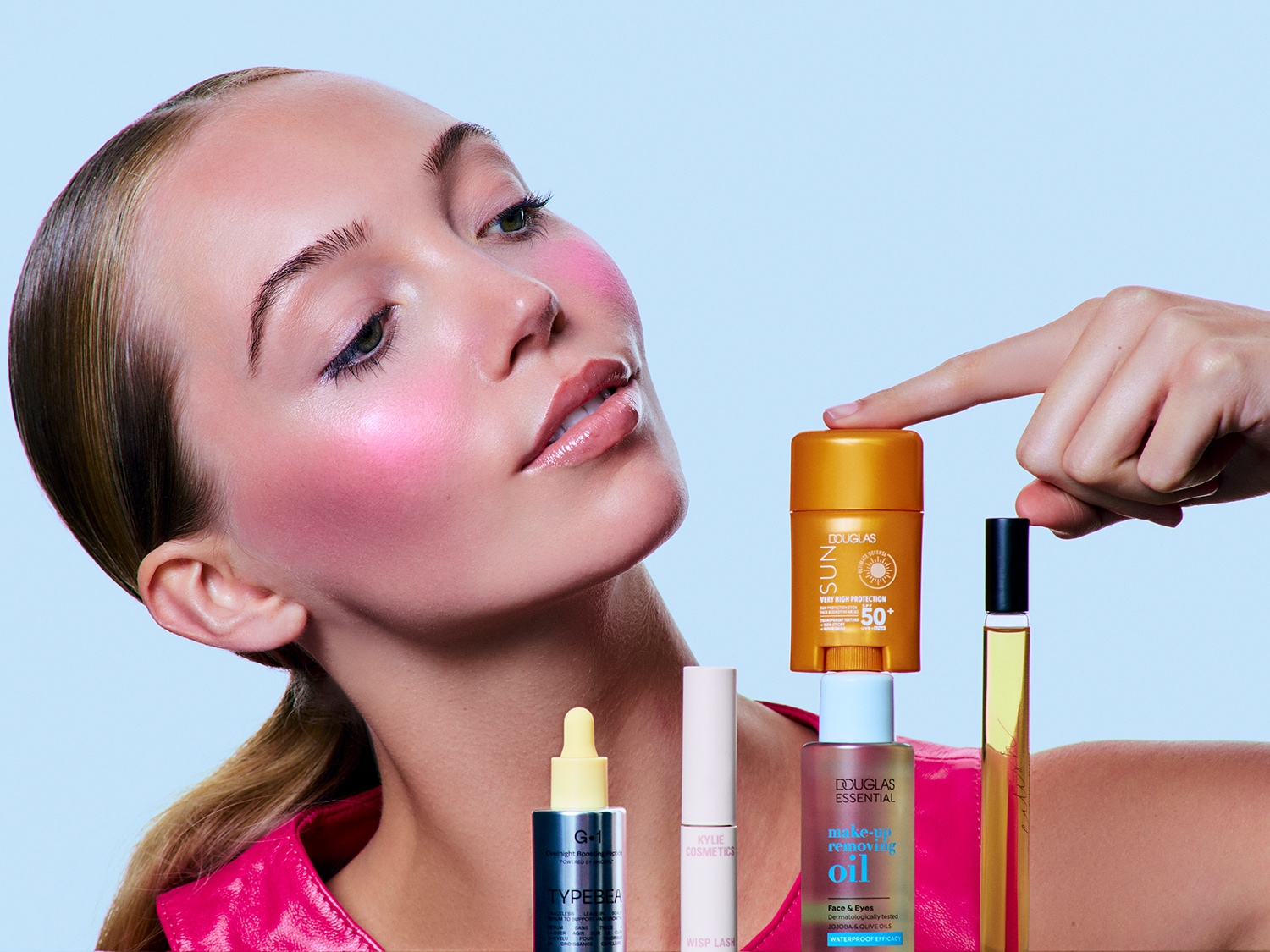 Ein Model mit rosa Rouge präsentiert eine Auswahl an Schönheitsprodukten, darunter Sonnenschutz von Douglas Sun, Make-up-Entferneröl von Douglas Essential und Wimperntusche von Kylie Cosmetics.