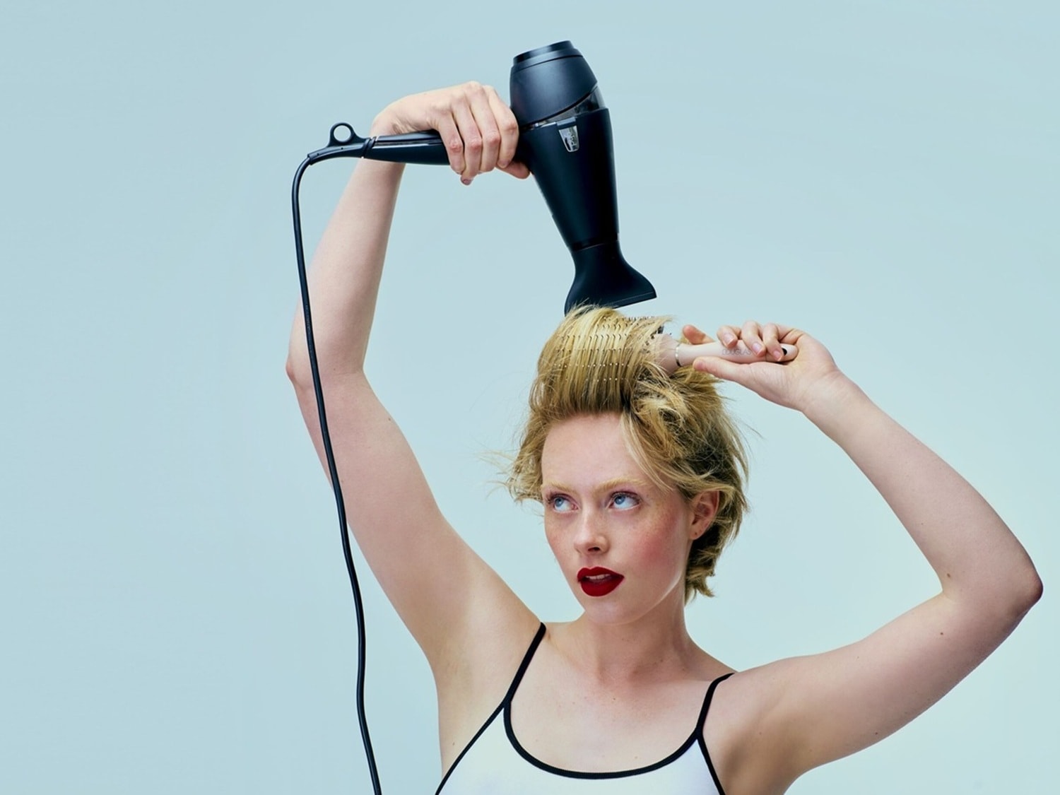 A woman with red lipstick and fair skin holds a black hair dryer above her head, styling her short blonde hair with a round brush.