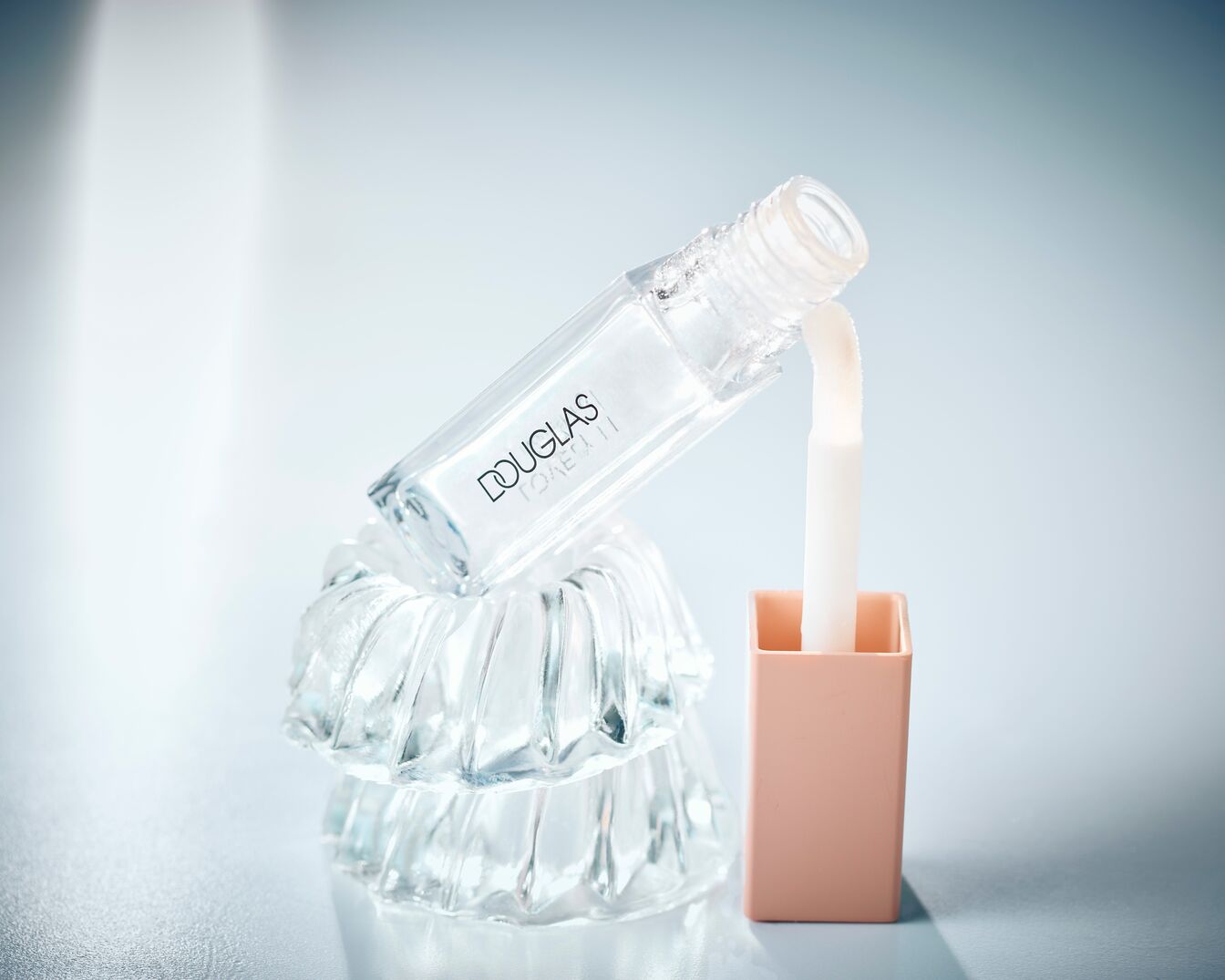 Klarer Douglas Lipgloss in transparenter Verpackung mit rosafarbenem Deckel, der auf einem Glasornament steht.