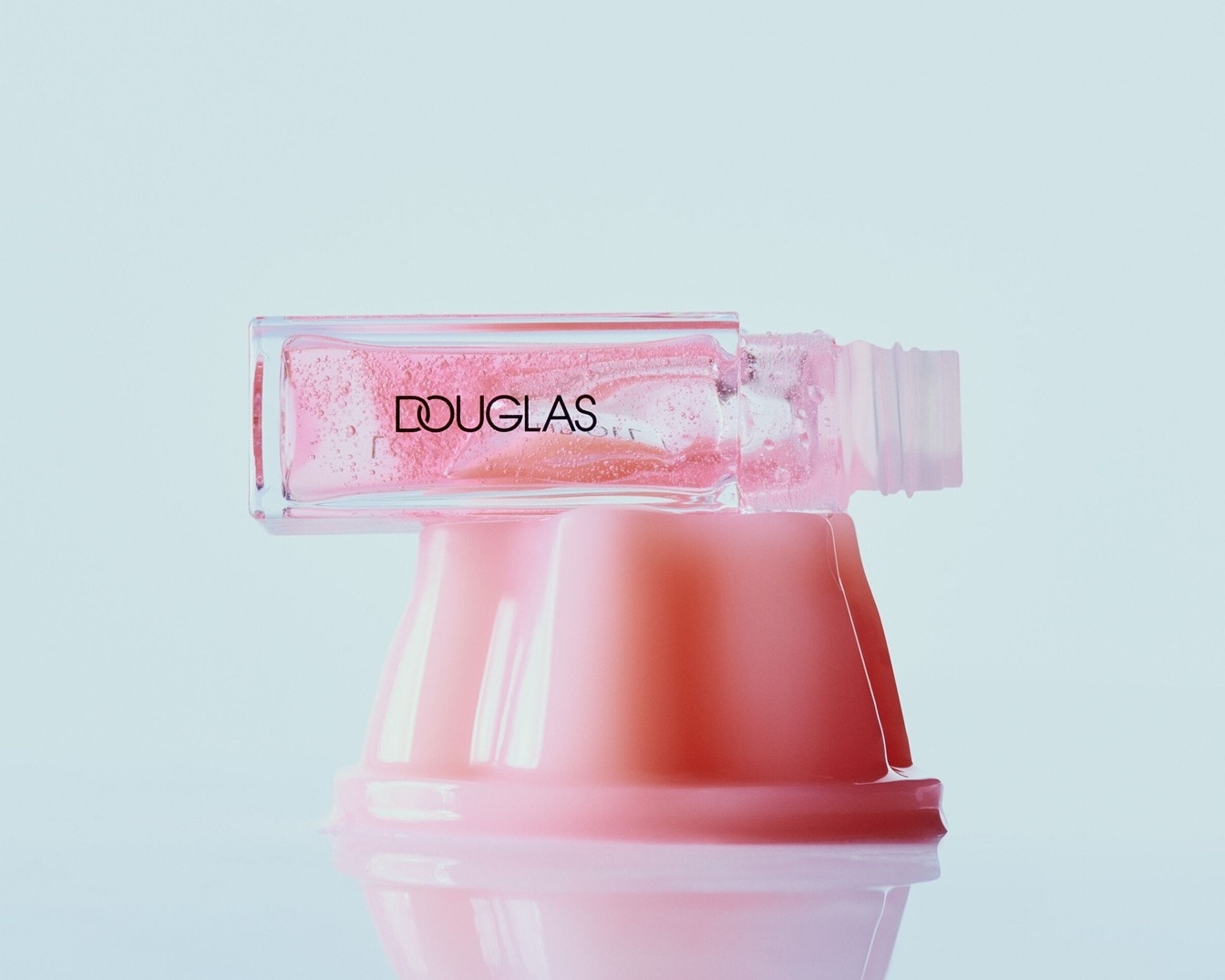 Das Douglas Lipgloss präsentiert sich in einer durchsichtigen Verpackung mit rosafarbenem Inhalt, der auf einem rosa Podest liegt.
