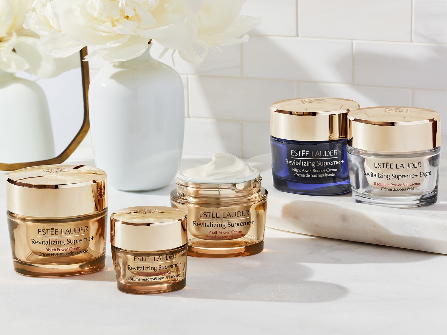 Fünf Tiegel verschiedener Revitalizing Supreme+ Cremes von Estée Lauder, darunter Youth Power Creme und Night Power Bounce Creme, präsentiert in eleganten goldenen und blauen Tiegeln auf marmoriertem Untergrund.