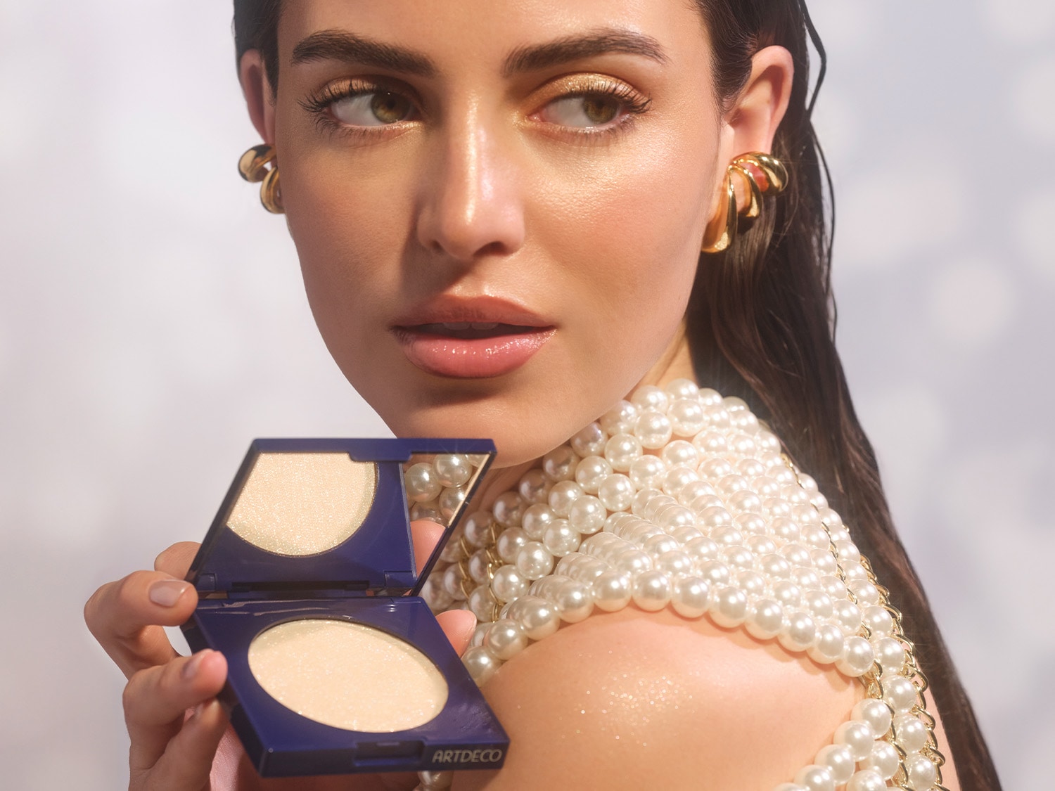 Eine Frau mit goldenem Lidschatten und einem Oberteil aus Perlen hält das Diamond Beauty Powder von ARTDECO in einem marineblauen Gehäuse.