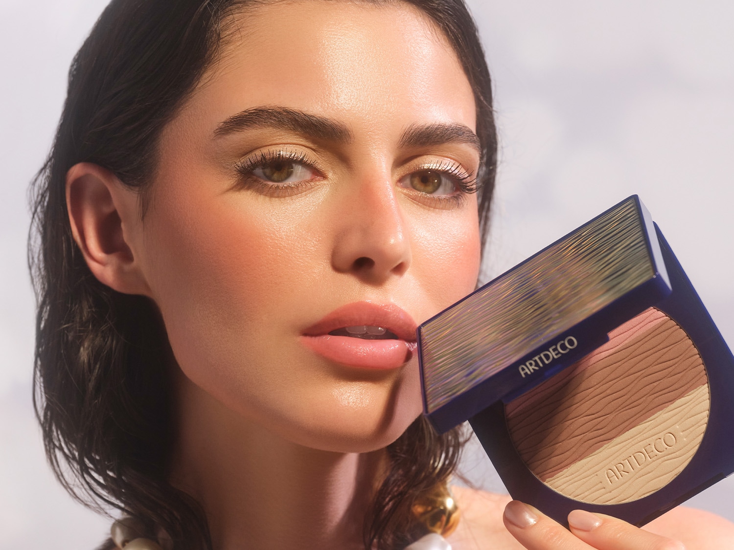 Nahaufnahme eines Models mit dem All Seasons Bronzing Powder von ARTDECO in der Hand, das in einem marineblauen Etui mit drei Farbtönen von hell nach dunkel präsentiert wird.