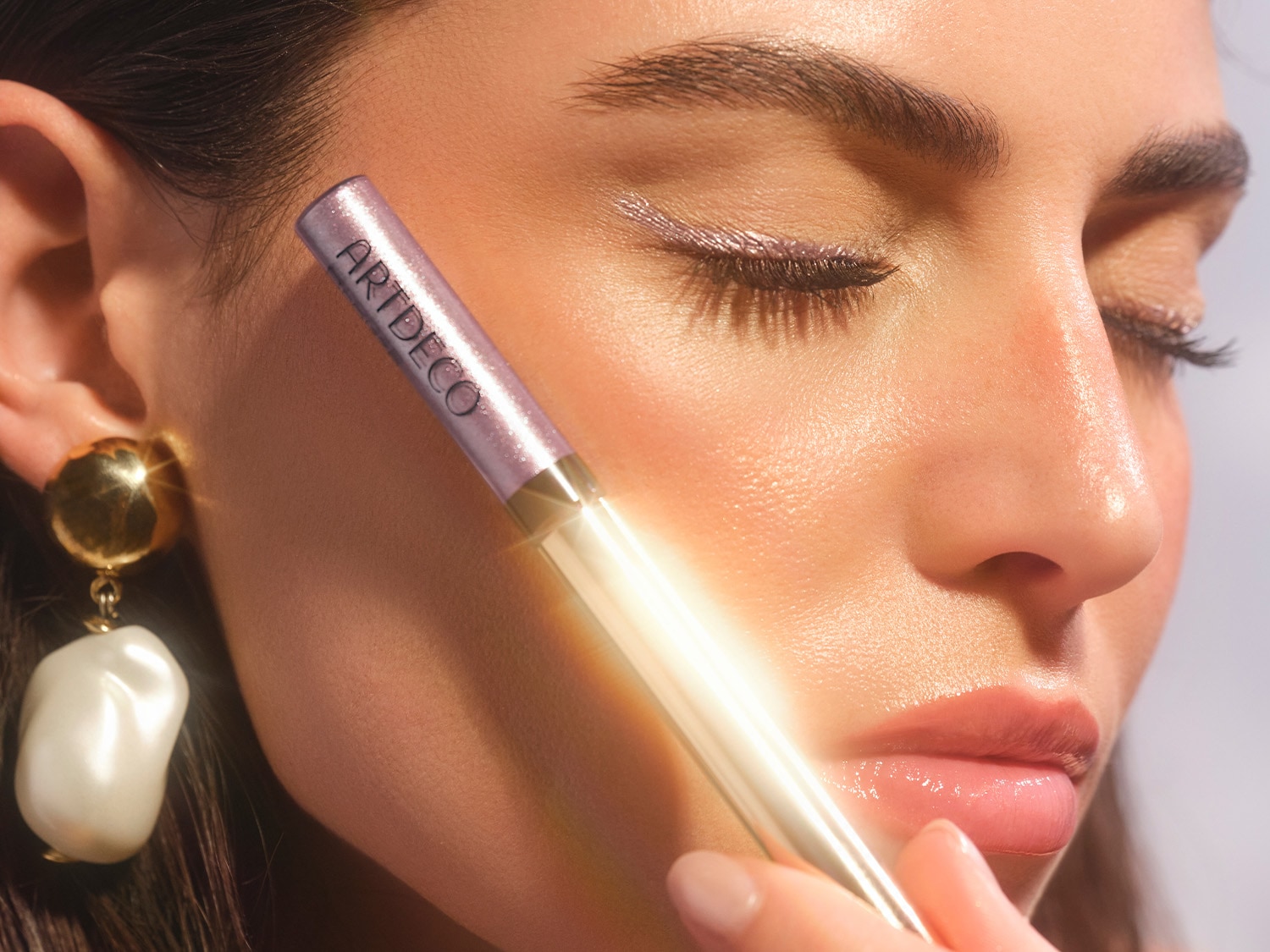 Nahaufnahme einer Frau, die mit einem lila schimmernden Eyeliner von ARTDECO einen Lidstrich gezogen hat, dazu trägst sie goldene Ohrringe mit einer weißen Perle.