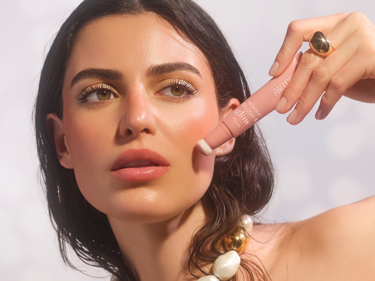Eine Frau mit heller strahlender Haut und Perlenkette hält eine Tube Wonder Blush & Glow von ARTDECO an ihre Wange.